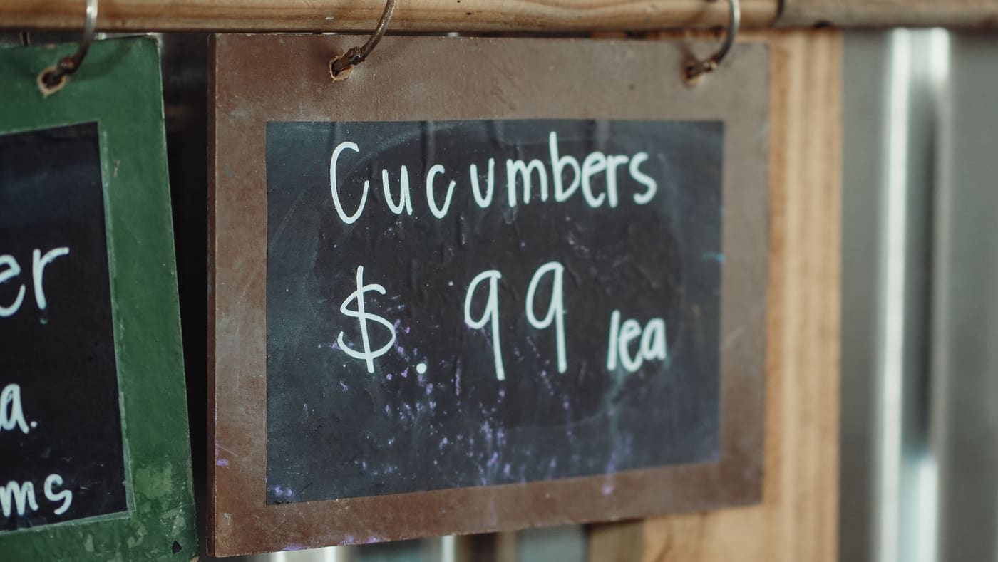 A small chalkboard sign showing a price for cucumbers hung on a wooden market stall