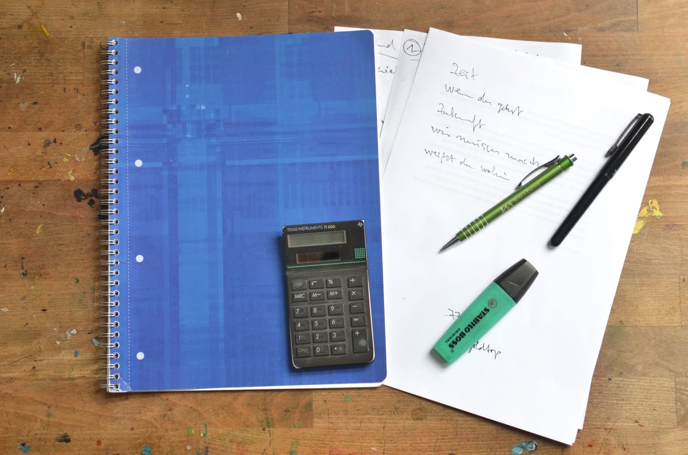 A blue spiral notebook, small calculator, highlighter, and handwritten papers on a wooden desk