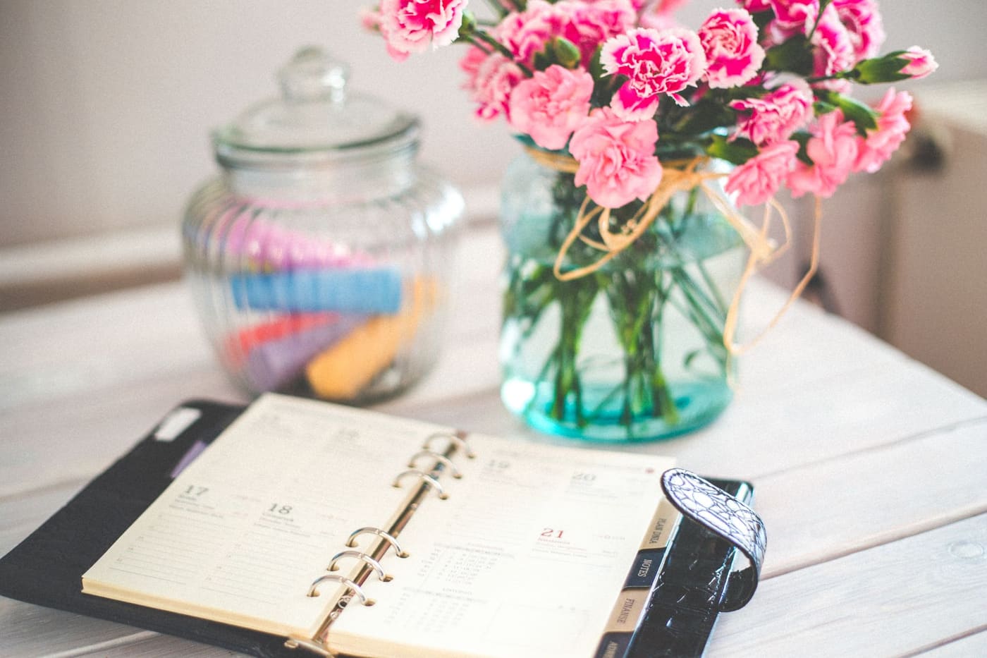 Open ring-bound planner on a white table beside a jar and a vase of pink flowers