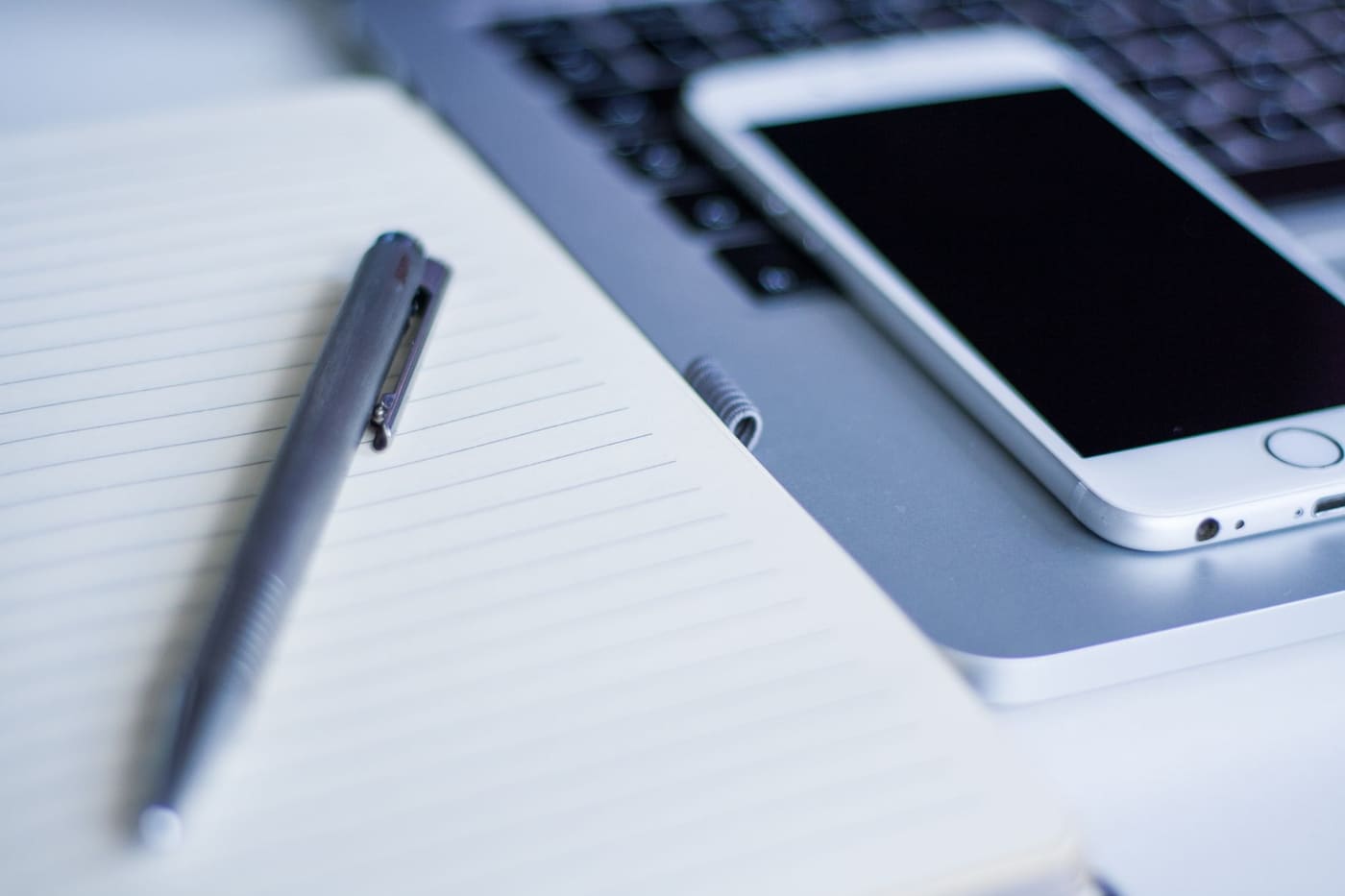 Open notebook with a pen beside a smartphone and part of a laptop on a desk