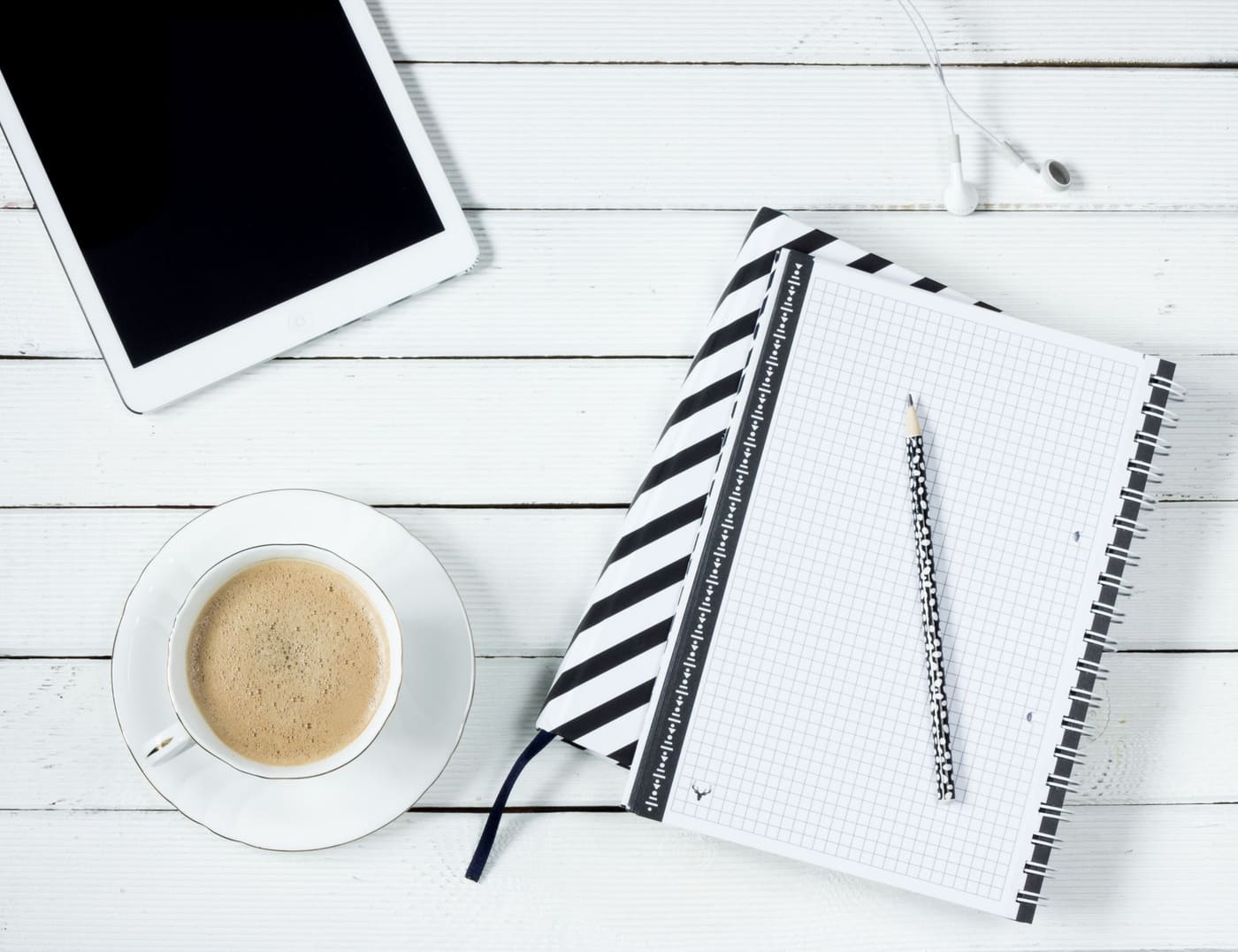 Flat lay of a tablet, a cup of coffee and a striped notebook with a pencil on a white table