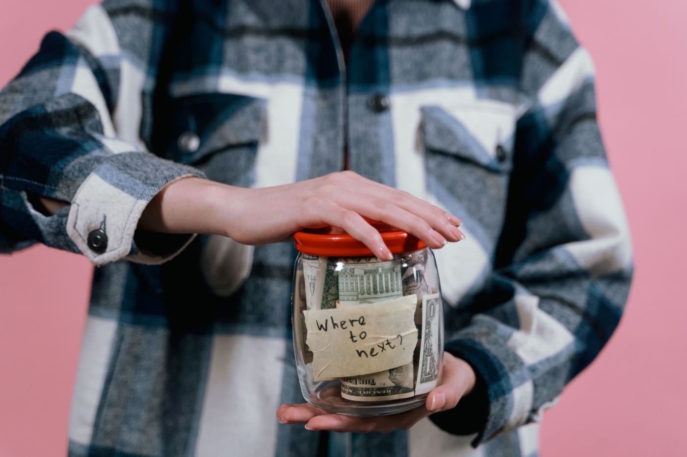 A person in a plaid shirt holding a glass jar of cash with a handwritten 'Where to next?' label