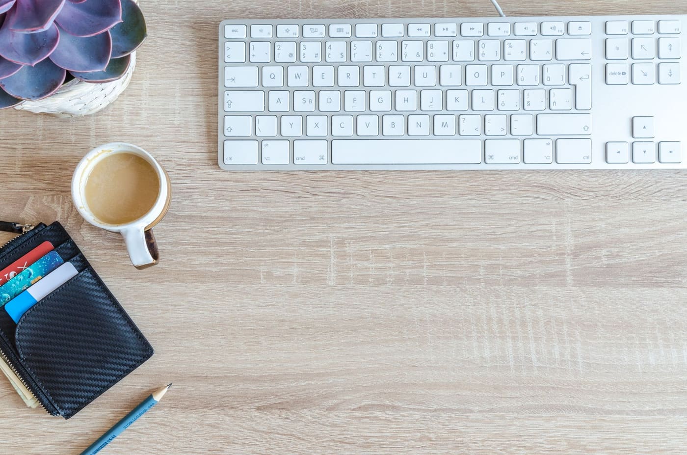 A small black wallet and a cup of coffee on a wooden desk beside a keyboard