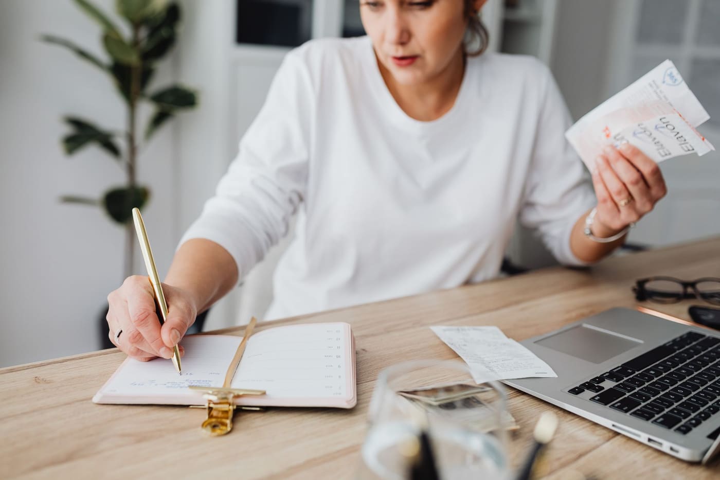 A woman at a desk writing in a notebook while holding a few receipts, with a laptop beside her